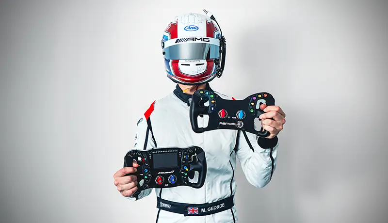 Matthew George as he dives into the development of our GT steering wheel, comparing its performance to the Ascher Racing Artura Sim wheel range. Matt is holding both race wheels to show the similarities. He is wearing his white race suit, wearing a helmet with the visor down. Photographed on a white background